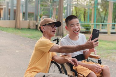 Şehir parkında tekerlekli sandalyede oturan baba ve özürlü çocuklar akıllı telefondan selfie çekerek, özel çocukların eğitim çağında hayatlarıyla, aile tatili konseptinde mutlu engelli çocuklarla seyahat ederek eğleniyorlar..