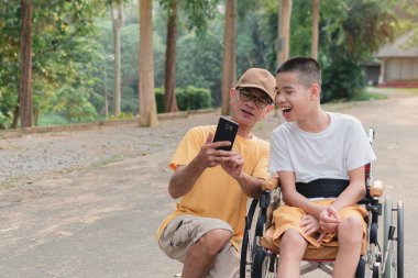 Şehir parkında tekerlekli sandalyede oturan baba ve özürlü çocuklar akıllı telefondan selfie çekerek, özel çocukların eğitim çağında hayatlarıyla, aile tatili konseptinde mutlu engelli çocuklarla seyahat ederek eğleniyorlar..