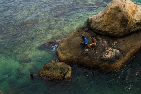 Dos chicos locales sentados en la roca rodeados de agua otro nadando — Foto de Stock