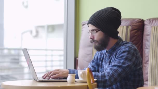 Joven hombre de moda hipster trabajando en el ordenador portátil en la cafetería — Vídeos de Stock