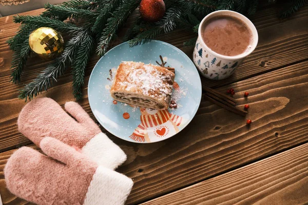 Primer plano de un strudel en un plato de Navidad cerca de manoplas y chocolate caliente en una mesa de madera. Desayuno de Navidad —  Fotos de Stock