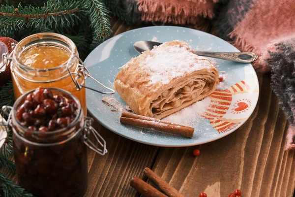 Closeup de um strudel em uma chapa de Natal perto de paus de canela e uns jarros de engarrafamento. Café da manhã de Natal em uma mesa de madeira — Fotografia de Stock