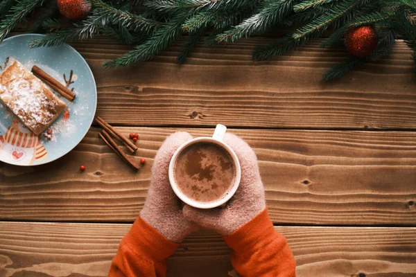 Primeros planos con manoplas sosteniendo una taza de chocolate caliente sobre un fondo de madera con strudel. Bebida caliente de Navidad —  Fotos de Stock