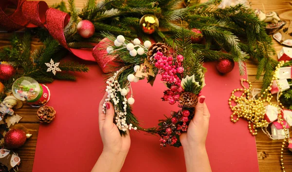 Hands making rustic Christmas wreath s on a red background. Atmospheric moody image at holiday workshop — Stock Photo, Image