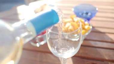 Yaz bahçesindeki bir bardağa beyaz şarap dökülüyor. Yakın plan. Erkek eli. Işıklar ve sığ alan derinliği.