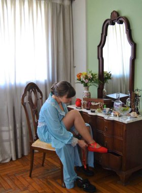 Mavi bornozlu kadın tuvalet masasında oturuyor ve kırmızı bale ayakkabılarını giyiyor.