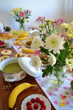 Üzerinde pembe benekli beyaz bir masa örtüsü olan bir kahvaltı masası ve üzerinde çiçekler, turta, meyve, bitkisel çay, çikolata, köfte, pirinç keki ve diğer lezzetli yemekler var.