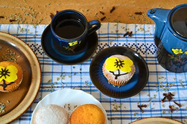 Blue coffee set with yellow flowers surrounded by muffins decorated with powdered sugar and icing in the shape of a yellow flower that matches those of the coffee set on a white and blue squares
