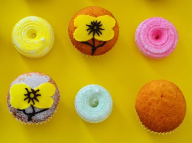 Pudra şekeriyle süslenmiş sıra sıra muffinler ve sarı arkaplanda krema şeklinde sarı çiçekler.