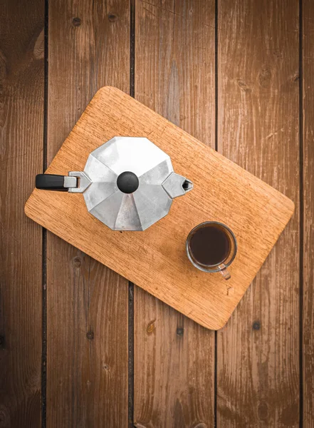 Metal moka demliğinin yanındaki şeffaf cam bardakta taze demlenmiş kahve. Yüzü gayzerli kahve makinesi ve ahşap arka planda şeffaf cam..