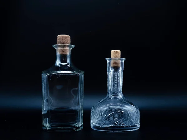 Empty glass carafe and empty glass bottle closed with cork caps isolated on a black background. Pair of the transparent bottles. Front view of the two vertical staying jars with different shape.