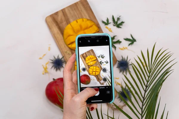 Female hand with a mobile taking a photo of a mango on a wooden table from above with some flowers and a tropical leaf