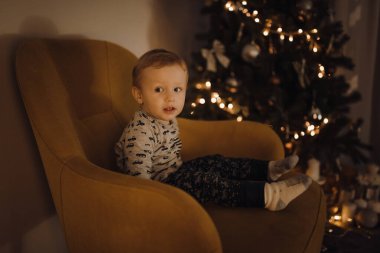 Mutlu çocuk arka planda Noel ağacı ve süslemeleri olan bir koltukta oturuyor..