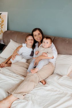 Anne ve iki oğlu birlikte bir yatakta otururken rahatlıyor ve gülümsüyorlar.