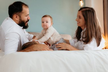 Bebekli ebeveynler beyaz bir yatakta vakit geçirip kaynaşıyorlar.