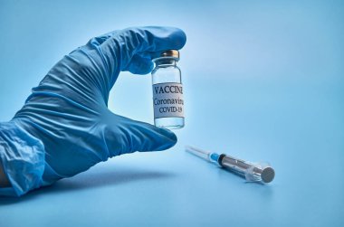 Bir şişe COVID-19 aşısı ve doktorlarda koronavirüs enfeksiyonuna karşı enjeksiyon iğnesi olan bir şırınga mavi arka planda bir nitrile eldiveni teslim ediyor. Yakın plan. Kopyalanacak yer. Konsept: