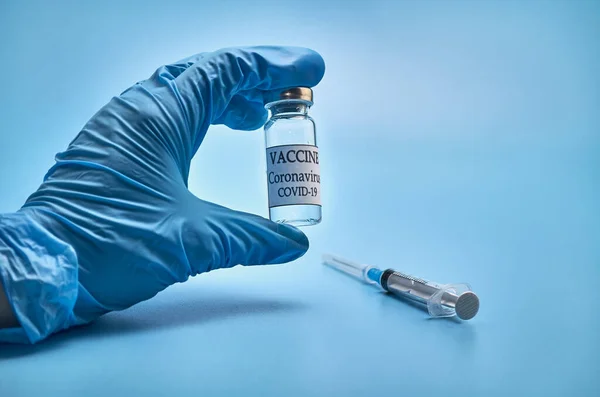 A bottle of COVID-19 vaccine and a syringe with an injection needle against coronavirus infection in the doctors hand in a nitrile glove on a blue background. Close-up. Place to copy. The concept of