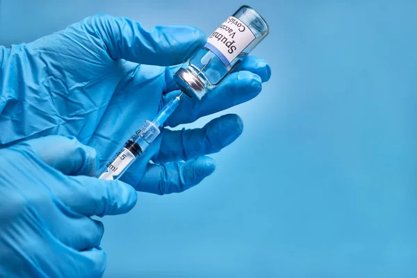 Lahvička vakcíny COVID-19 Sputnik V a injekční stříkačka s injekční jehlou proti infekci koronavirem v rukou lékaře v nitrilové rukavici na modrém pozadí. Detailní záběr. Místo na kopírování. The Stock Obrázky