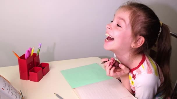 Skolflicka, liten brunett flicka gör lektioner i vardagsrummet vid bordet. Flickan skrattar. På bordet finns en anteckningsbok, en lärobok, pennor, skolmaterial. Ryssland. — Stockvideo