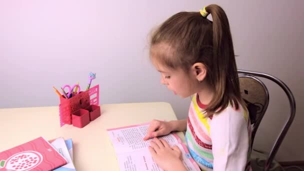 Skolflicka, liten brunett flicka gör lektioner i vardagsrummet vid bordet. På bordet finns en anteckningsbok, en lärobok, pennor, skolmaterial. Ryssland. — Stockvideo