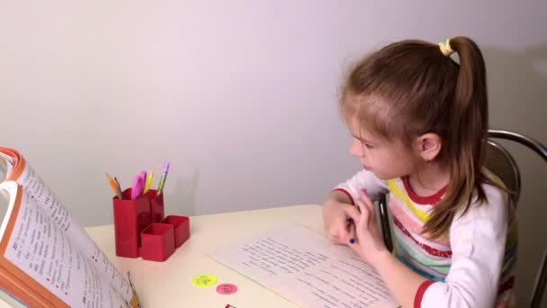 Schoolmeisje, klein brunette meisje doet lessen in de woonkamer aan tafel. Op tafel ligt een notitieboekje, een handboek, pennen, schoolspullen. Rusland. — Stockvideo