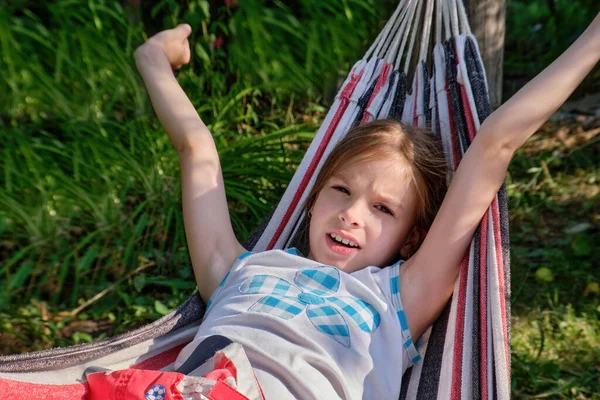 Una niña feliz se encuentra en una hamaca en el jardín en el verano. Un niño en una hamaca durante las vacaciones sonríe en el parque contra el fondo de hierba verde. —  Fotos de Stock