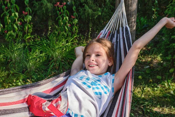 Una niña feliz se encuentra en una hamaca en el jardín en el verano. Un niño en una hamaca durante las vacaciones sonríe en el parque contra el fondo de hierba verde. —  Fotos de Stock