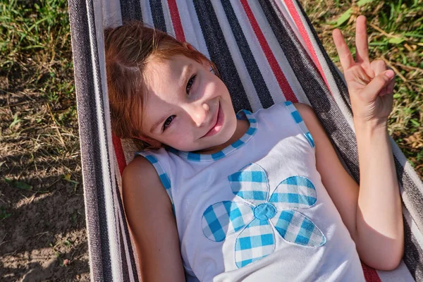 Una niña feliz se encuentra en una hamaca en el jardín en el verano. Un niño en una hamaca durante las vacaciones sonríe en el parque contra el fondo de hierba verde. —  Fotos de Stock