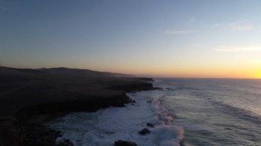 Kıyı şeridinde gün batımında kayalıkları, renkleri ve deniz fark ediliyor.
