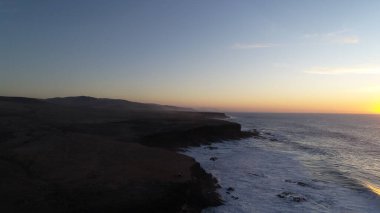 Kıyı şeridinde gün batımında kayalıkları, renkleri ve deniz fark ediliyor.