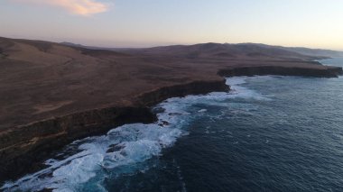 Kıyı şeridinde gün batımında kayalıkları, renkleri ve deniz fark ediliyor.