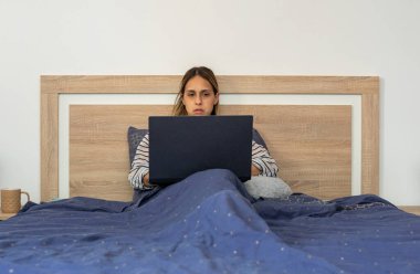 Genç bir kadın, otuzlu yaşlarda, laptopuyla telgraf çekiyor, mavi bir yorgan altında, yatak odasının konforunda oturuyor..