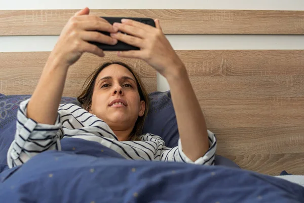 Genç beyaz kadın, cep telefonuyla video görüşmesi yapıyor, telefondaki kameraya bakıyor, yatak odasındaki konforlu yatağında gülümsüyor ve merhaba diyor..