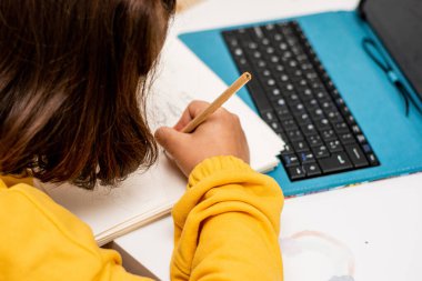 Bir elinde alçı olan, sarı kazaklı, eskiz defterine, yatak odası masasına çizim malzemeleri ve tabletle çizim yapan bir kızın detaylı fotoğrafı..