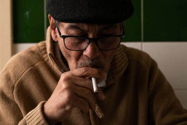 Beyazlı, bej kazak giyen, koyu gri bereli, siyah gözlüklü, ağzında sigara içen düşünceli bir adamın yere bakışları..