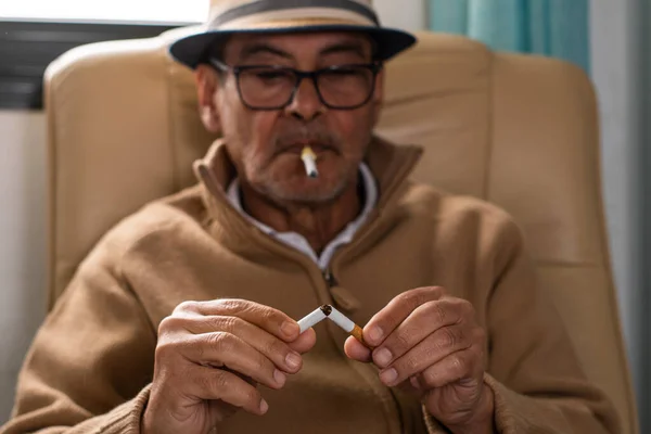 Elderly Caucasian man wearing a hat and black glasses, holding in his mouth a plastic cigarette to stop smoking, and in his hands a cigar that he has broken who looks thoughtful as a sign of rejection of tobacco