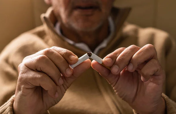 Unrecognizable senior man, out of focus with his hands in focus holding a broken cigarette in his hands looking thoughtful, thinking about quitting smoking.