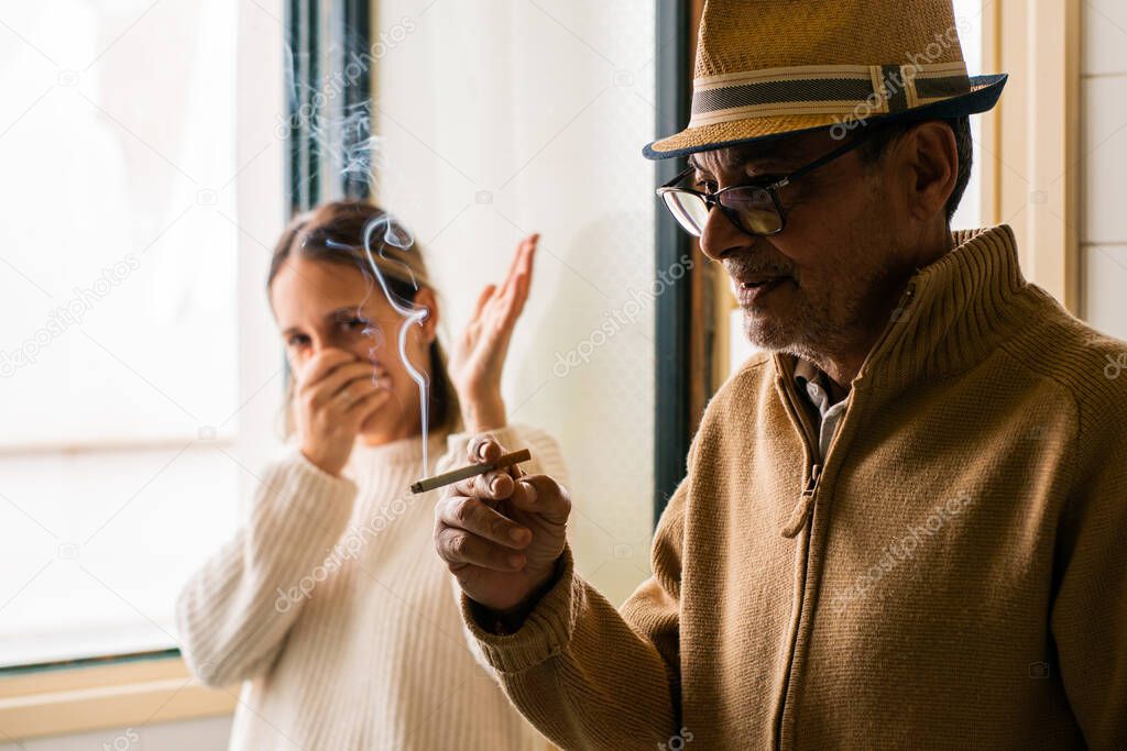 Elderly, Caucasian man, in the foreground, smoking a cigarette oblivious to the discomfort that the smoke produces on a young, Caucasian girl, who is in the background out of focus, with disapproving gestures.