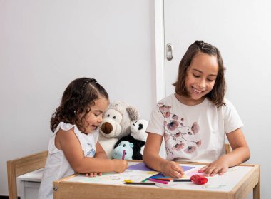 İki beyaz kız, çok gülümsüyorlar, çocuk masasında oturuyorlar, babaları için kartpostal ve kartpostal çiziyorlar..