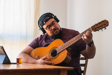 Beyaz, iri yarı, sakallı, gözlüklü ve şapkalı bir adam. Tabletinizden online gitar dersi alınıyor.