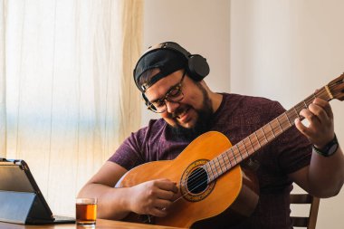 Beyaz, iri yarı, sakallı, gözlüklü ve şapkalı bir adam. Tabletinizden online gitar dersi alınıyor.