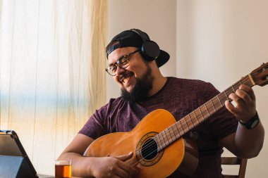 Beyaz, iri yarı, sakallı, gözlüklü ve şapkalı bir adam. Tabletinizden online gitar dersi alınıyor.