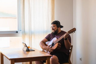 Beyaz, iri yarı, sakallı, gözlüklü ve şapkalı bir adam. Tabletinizden online gitar dersi alınıyor.