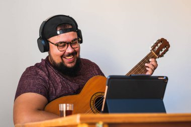 Şişman, sakallı, gözlüklü, kulaklıklı, şapkalı, gülümseyen, tabletinden online gitar dersi alan bir adam..