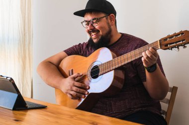 Şişman, sakallı, gözlüklü, kulaklıklı, şapkalı, gülümseyen, tabletinden online gitar dersi alan bir adam..