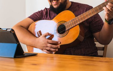Tanınmaz halde sakallı bir adam. Tabletinden online gitar dersi aldığı için gülümsüyor. Evdeki oturma odasında oturuyor..