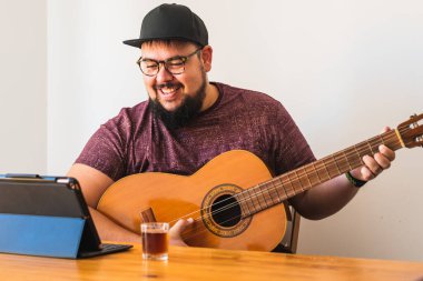 Şişman, sakallı, gözlüklü, kulaklıklı, şapkalı, gülümseyen, tabletinden online gitar dersi alan bir adam..