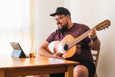 Şişman, sakallı, gözlüklü, kulaklıklı, şapkalı, gülümseyen, tabletinden online gitar dersi alan bir adam..