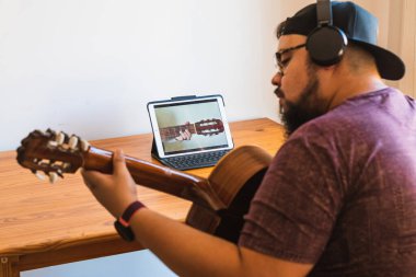Şişman, sakallı, gözlüklü, kulaklıklı, şapkalı, gülümseyen, tabletinden online gitar dersi alan bir adam..