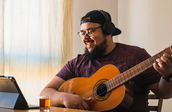 Beyaz, iri yarı, sakallı, gözlüklü ve şapkalı bir adam. Tabletinizden online gitar dersi alınıyor.